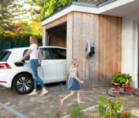 Ein weißes E-Auto steht halb in einer mit Holz verkleideten Garage an einem weißen Haus; eine Frau steckt das Kabel der Wallbox ins Auto. Ein Kind läuft vorbei ins Haus..