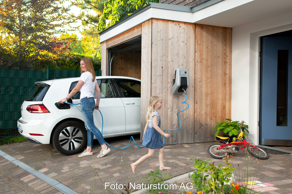 Ein weißes E-Auto steht halb in einer mit Holz verkleideten Garage an einem weißen Haus; eine Frau steckt das Kabel der Wallbox ins Auto. Ein Kind läuft vorbei ins Haus..