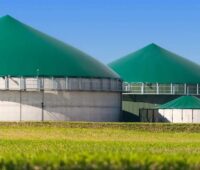 Hinter einen kurz gemähten, grünen Wiese liegt eine Biogasanlage mit zwei Fermentern vor einem blauen Himmel.