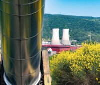 Blick auf ein Geothermie-Krafterk in Larderello. Im Vordergrund eine dicke Fernwärmeleitung und blühender Ginster.