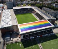 Eine PV-Anlage in Regenbogenfarben auf dem Dach einer von vier überdachten Tribünen eines Fussballstadions.