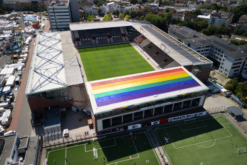 Eine PV-Anlage in Regenbogenfarben auf dem Dach einer von vier überdachten Tribünen eines Fussballstadions.
