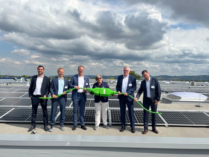 6 Personen auf einem Industriedach mit Photovoltaik mit symbolischem Stecker.