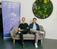 Zwei Männer in legerer Kleidung auf einem Sofa im Büroumfeld.