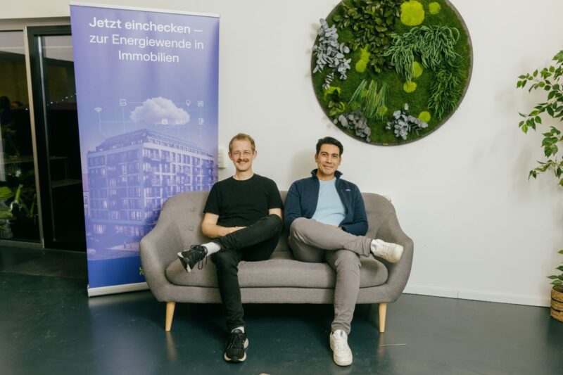 Zwei Männer in legerer Kleidung auf einem Sofa im Büroumfeld.