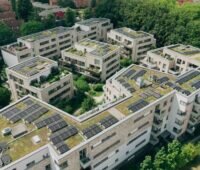 Luftaufnahme einer mehrteiligen Wohnanlage mit begrünten Flachdächern und zahlreichen Photovoltaikmodulen. Die Solaranlagen sind auf mehreren Gebäuden installiert und von grünen Bäumen umgeben.