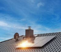 Mehrere schwarze Solarmodule sind auf dem geneigten Ziegeldach eines Einfamilienhauses installiert. Im Hintergrund sind ein Schornstein, eine Satellitenschüssel und ein blauer Himmel mit Wolken zu sehen. Sonnenlicht spiegelt sich auf einem der Module.