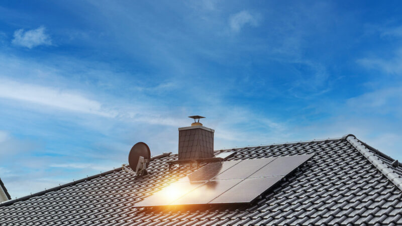 Mehrere schwarze Solarmodule sind auf dem geneigten Ziegeldach eines Einfamilienhauses installiert. Im Hintergrund sind ein Schornstein, eine Satellitenschüssel und ein blauer Himmel mit Wolken zu sehen. Sonnenlicht spiegelt sich auf einem der Module.