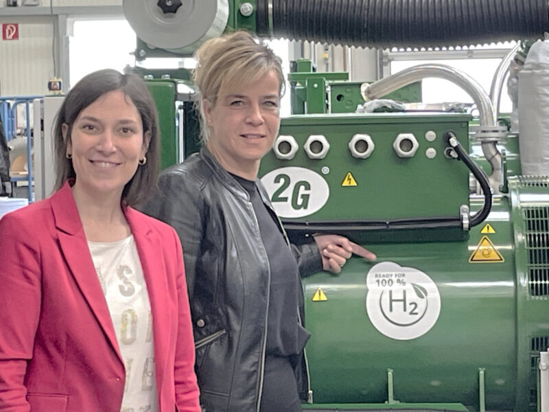 Zwei Frauen, Heike Wermer und Mona Neubaur, vor einem BHKW. Neubur zeigt auf einen Aufkleber mit der Aufschrift "Ready for 100 % H2"