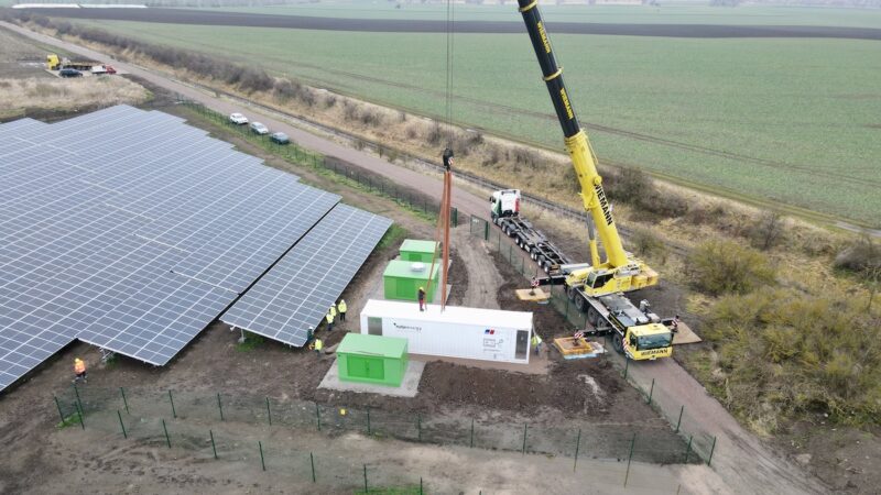 Zu sehen ist eine Luftaufnahme einer Photovoltaik-Freiflächenanlage mit Speicher in einem weißen Container, der gerade von einem Kran abgestellt wird.