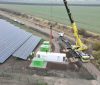 Zu sehen ist eine Luftaufnahme einer Photovoltaik-Freiflächenanlage mit Speicher in einem weißen Container, der gerade von einem Kran abgestellt wird.