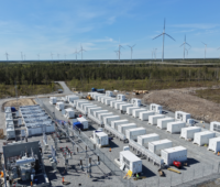 Batteriespeichersysteme auf Beton mit Umspannanlage auf einem Freigelände, im Hintergrund Windkraftanlagen.