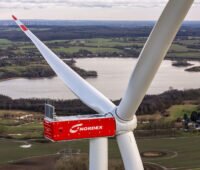 Luft-Nahaufnahme einer Windenergieanlage der Nordex Group mit roter Maschinenhausverkleidung und drei Rotorblättern. Im Hintergrund sind eine Seenlandschaft, Felder, Ortschaften und weitere Windenergieanlagen zu sehen.