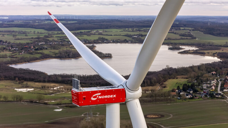 Luft-Nahaufnahme einer Windenergieanlage der Nordex Group mit roter Maschinenhausverkleidung und drei Rotorblättern. Im Hintergrund sind eine Seenlandschaft, Felder, Ortschaften und weitere Windenergieanlagen zu sehen.