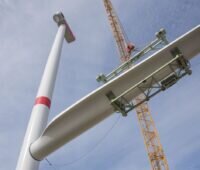 Rechts der Mast einer großen Windkraftanlage, ein Kran transportiert einen Flügel in die Höhe. Windenergieausbau