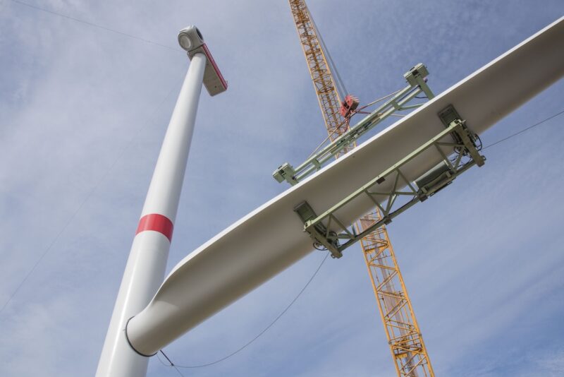 Rechts der Mast einer großen Windkraftanlage, ein Kran transportiert einen Flügel in die Höhe. Windenergieausbau