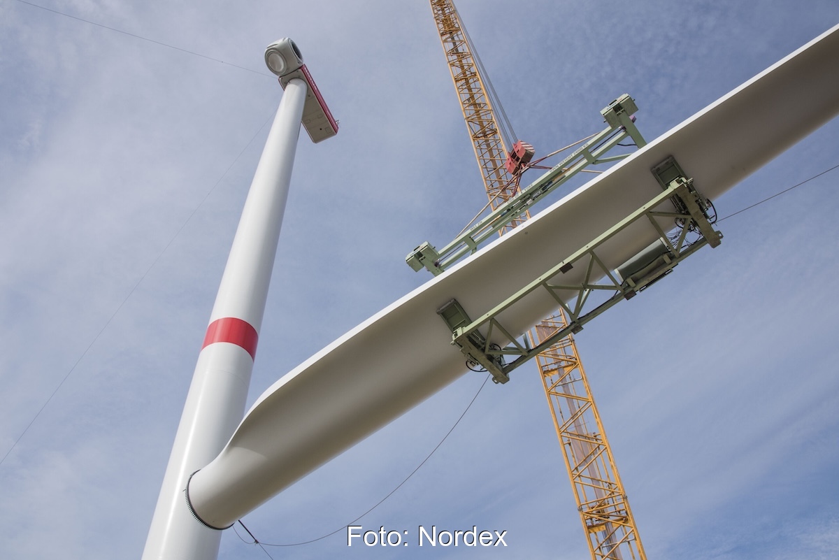 Rechts der Mast einer großen Windkraftanlage, ein Kran transportiert einen Flügel in die Höhe. Windenergieausbau