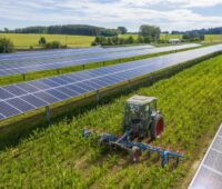 Aufgeständerte Solarmodulreihen beschatten eine Agrarfläche, dazwischen fährt ein Traktor.