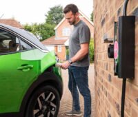 Ein Mann schließt ein grünes Elektroauto an eine Wallbox an einer Hauswand an.
