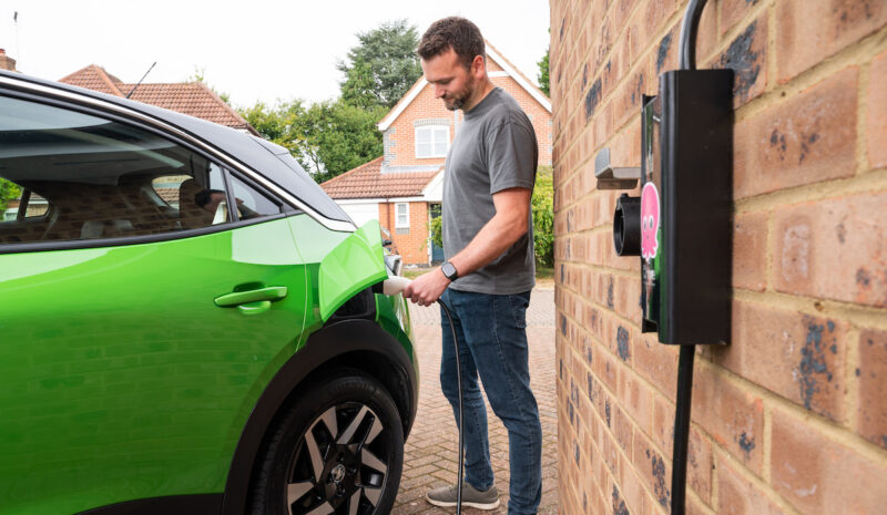 Ein Mann schließt ein grünes Elektroauto an eine Wallbox an einer Hauswand an.