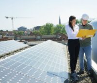 Eine Frau und ein Mann stehen auf einem Flachdach mit PV-Modulen und studieren Unterlagen.