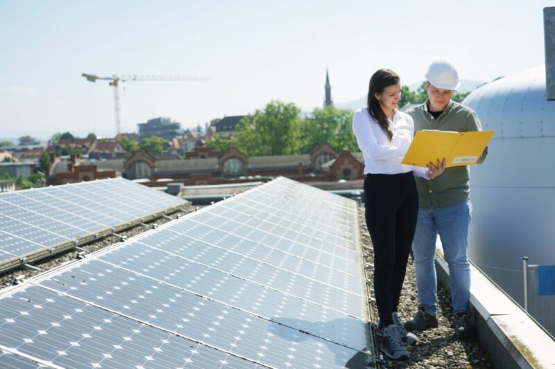 Eine Frau und ein Mann stehen auf einem Flachdach mit PV-Modulen und studieren Unterlagen.