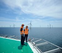 Zwei Mitarbeiter in Sicherheitswesten auf einem Hubschrauberlandeplatz einer Offshore-Plattform vor Windanlagen im Meer.