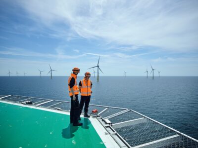 Zwei Mitarbeiter in Sicherheitswesten auf einem Hubschrauberlandeplatz einer Offshore-Plattform vor Windanlagen im Meer.