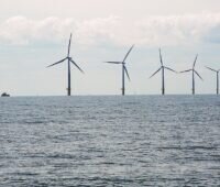 Fünf Windenergieanlagen stehen in einer Reihe im Meer; links fährt ein Schiff unter bewölktem Himmel. Grau-blaue Farben dominieren.