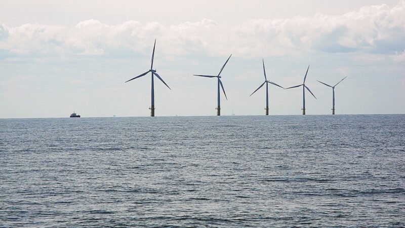 Fünf Windenergieanlagen stehen in einer Reihe im Meer; links fährt ein Schiff unter bewölktem Himmel. Grau-blaue Farben dominieren.