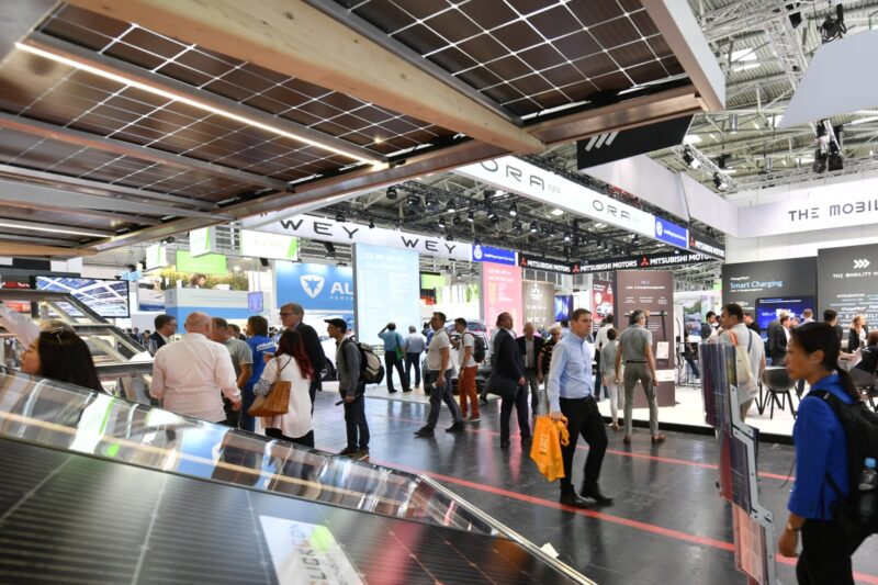 Passanten auf einer Messe mit Automobilständen und Photovoltaik.