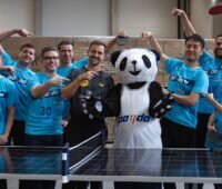 Timo Boll mit Stoff-Panda und Firmenvertreter:innen an einem als Tischtennisplatte aufgebauten Photovoltaik-Modul.