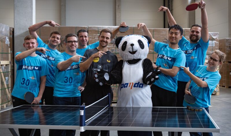 Timo Boll mit Stoff-Panda und Firmenvertreter:innen an einem als Tischtennisplatte aufgebauten Photovoltaik-Modul.
