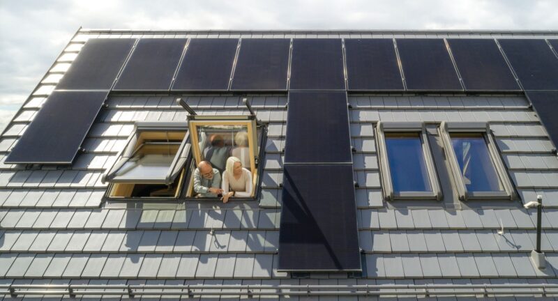 Zwei Menschen schauen aus Dachfenster zwischen Photovoltaikmodulen, Energy Sharing ist eine Alternative zur Einspeisevergütung von Solarstrom.