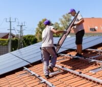 Zwei Installateure tragen ein Solarmodul auf das Dach, 1Komma5° hat ermittelt, welche Auswirkungen die PV-Strom-Direktvermarktung auf den Erlös der Verbraucher:innen hätte.