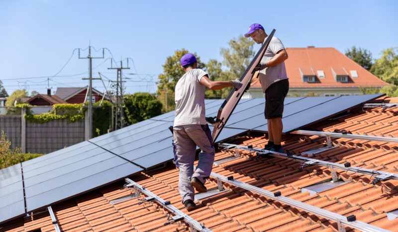 Zwei Installateure tragen ein Solarmodul auf das Dach, 1Komma5° hat ermittelt, welche Auswirkungen die PV-Strom-Direktvermarktung auf den Erlös der Verbraucher:innen hätte.