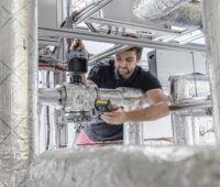 Ein Forscher zwischen dicken, mit Alufolie umwickelten Rohren, das Fraunhofer ISE optimiert Wärmepumpen für Industrieprozesse und Wärmenetze.