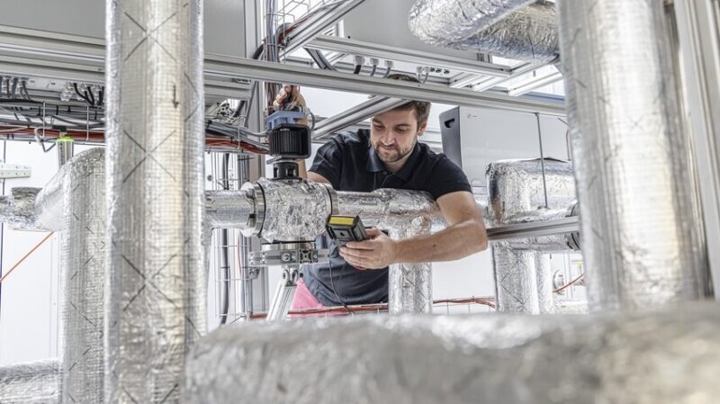 Ein Forscher zwischen dicken, mit Alufolie umwickelten Rohren, das Fraunhofer ISE optimiert Wärmepumpen für Industrieprozesse und Wärmenetze.