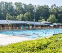 Schwimmbecken und im Hintergrund Gebäude mit Solarmodulen, das Freibad Hoheneck nutzt Solarstrom von der eigenen Photovoltaik-Anlage.