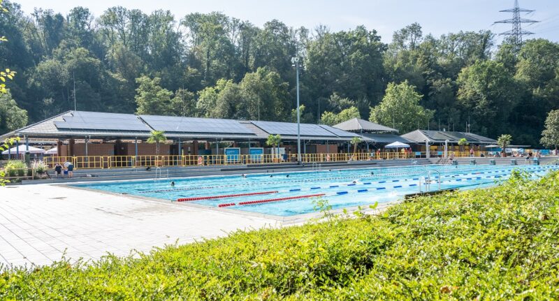 Schwimmbecken und im Hintergrund Gebäude mit Solarmodulen, das Freibad Hoheneck nutzt Solarstrom von der eigenen Photovoltaik-Anlage.