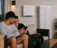 Ein Paar sitzt auf einen Sofa vor dem Haus neben dem Batteriespeicher Wall von Senec, den das Unternehmen auf der Intersolar/EES 2026 vorstellt.