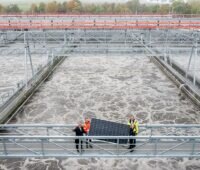 Offizielle zeigen ein Solarmodul auf einem Steg über dem Klärbecken, Stuttgarts größte Photovoltaik-Anlage entsteht als Faltdach über dem Hauptklärwerk.
