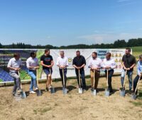 Spatenstich von 9 Personen auf Ackewrfläche vor Transparenten bei sonnigem Wetter.