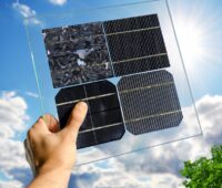 Eine Hand hält eine transparente Kunststoff-Platte mit vier verschiedenen Solarzellen vor blauem Himmel - Symbol für PV-Zellen-Technologien