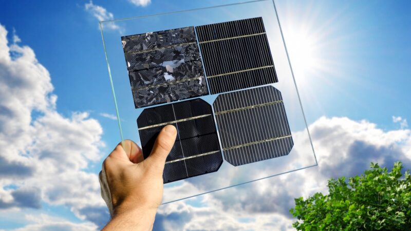 Eine Hand hält eine transparente Kunststoff-Platte mit vier verschiedenen Solarzellen vor blauem Himmel - Symbol für PV-Zellen-Technologien