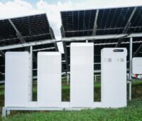 Vier weiße Batterieschränke und ein Wechselrichter stehen unter den aufgeständerten Modulen einer Photovoltaik-Freiflächenanlage.