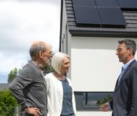 Ein älteres Ehepaar im Gespräch mit einem Herrn im Anzug vor ihrem Haus mit PV-Anlage auf dem Dach.