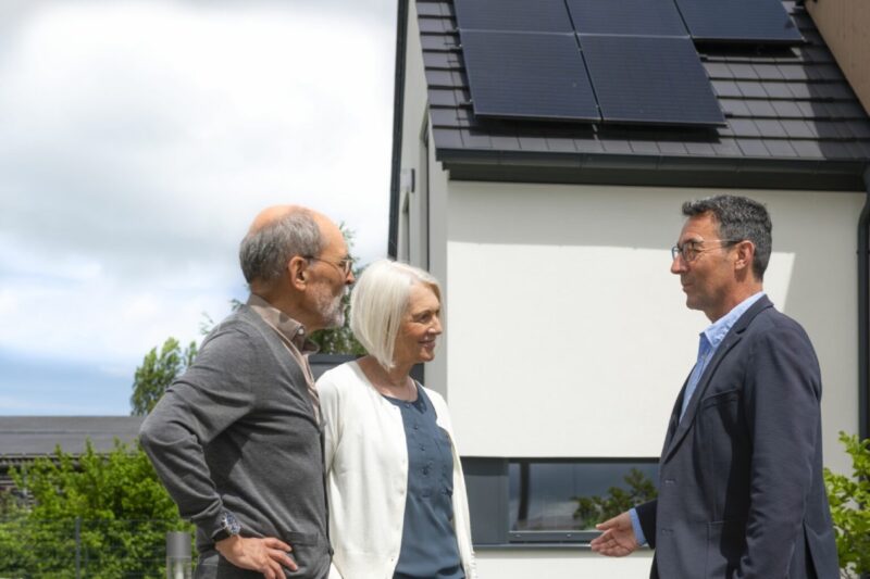 Ein älteres Ehepaar im Gespräch mit einem Herrn im Anzug vor ihrem Haus mit PV-Anlage auf dem Dach.