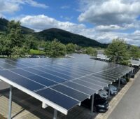 Überdachter Parkplatz mit Solarmodulen vor grünen Bäumen und Hügeln.