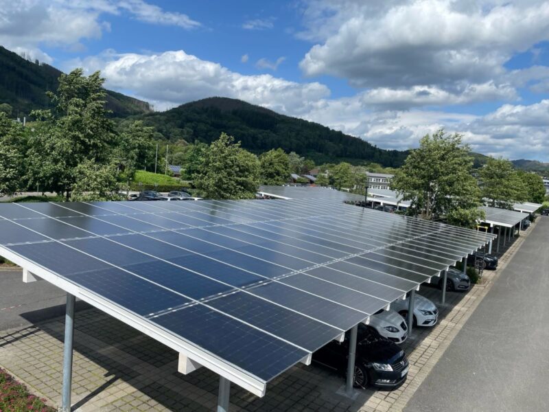 Überdachter Parkplatz mit Solarmodulen vor grünen Bäumen und Hügeln.